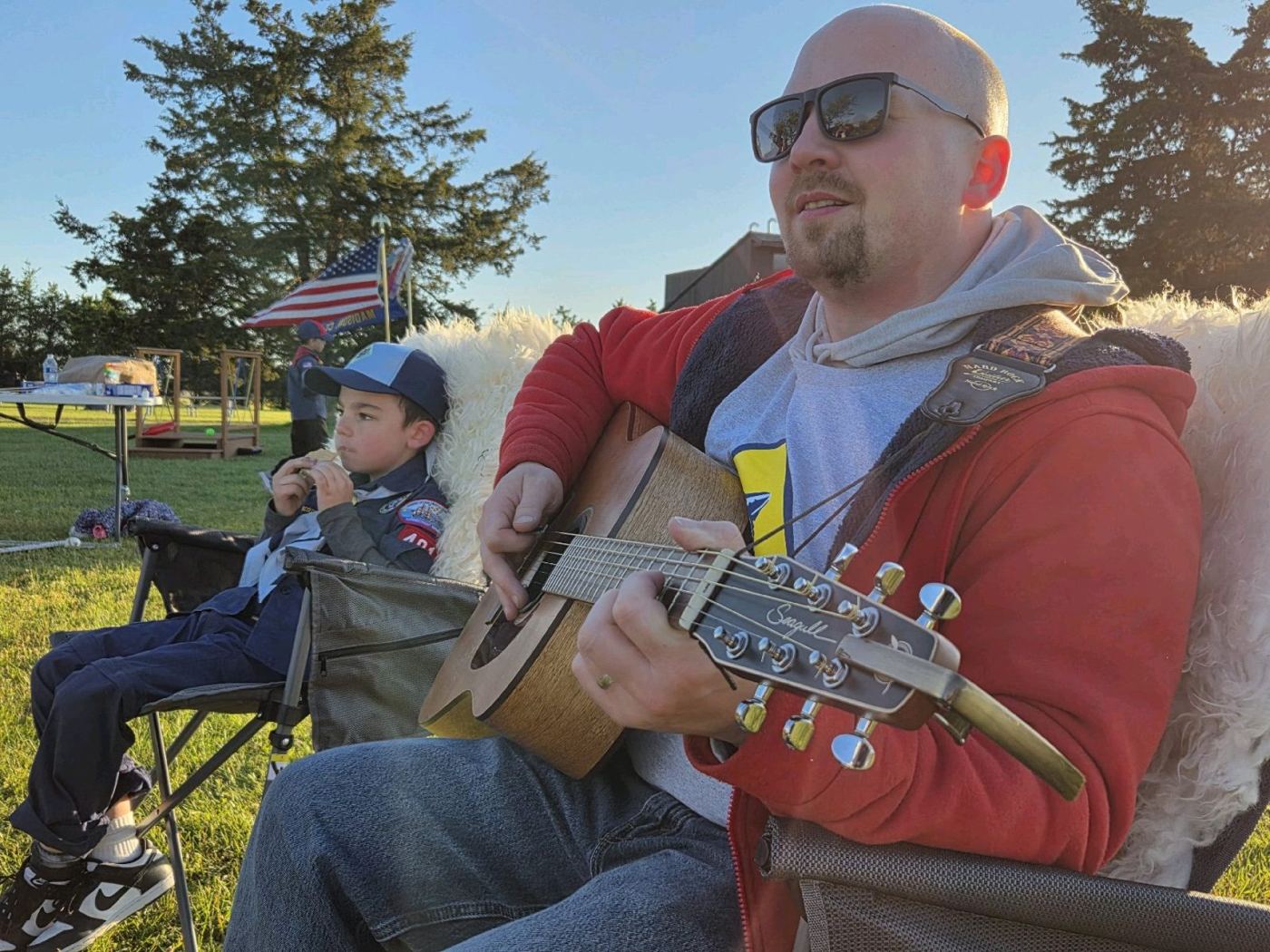A dad playing guitar next to a Cub Scout at camp